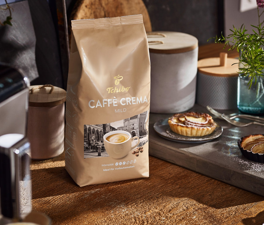 Sur une table en bois, un paquet de Caffè Crema Mild, une machine à café et des assiettes avec des gâteaux.