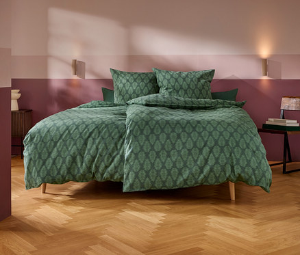 Une chambre à coucher avec un lit fait de linge de lit vert à motif de feuilles. Les murs sont peints en deux bandes horizontales, rose clair en haut et marron foncé en bas. Deux appliques murales éclairent l'espace. Une petite table de chevet en bois avec un objet décoratif blanc est à gauche, et une chaise en bois avec des livres est à droite. Le sol est en bois clair à chevrons.
