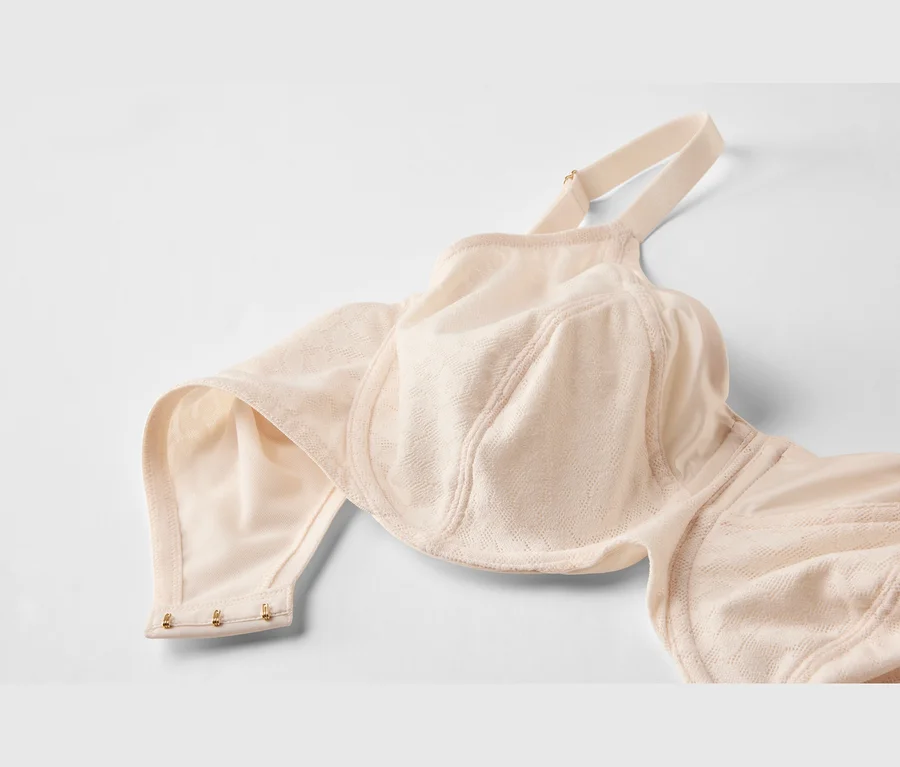 Un soutien-gorge beige clair avec des bonnets texturés et une bretelle visible est posé à plat. La fermeture à trois agrafes dorées est visible à gauche.