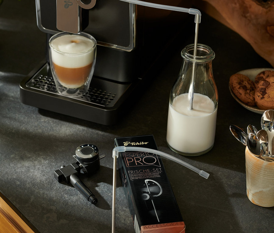 Une machine à café entièrement automatique Tchibo « Esperto Pro » anthracite prépare une boisson. Le lait passe par un tuyau dans une bouteille en verre. À côté se trouve un set fraîcheur Esperto Pro & Esperto2 Milk.