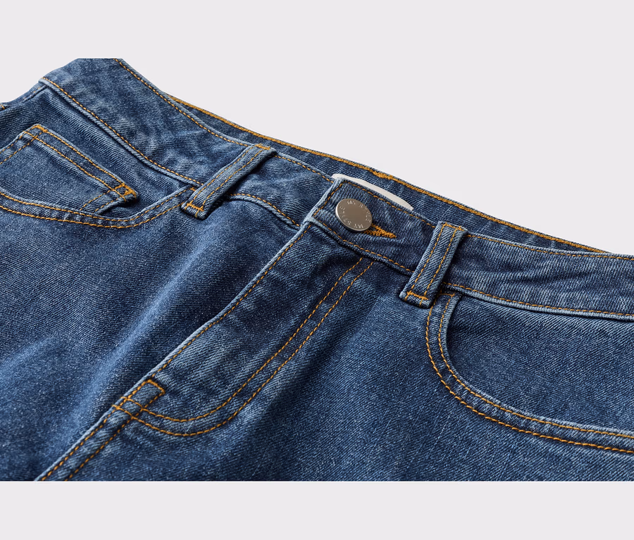 Gros plan sur la partie supérieure d'un jean en denim bleu, montrant la ceinture, les passants de ceinture, les poches avant et un bouton argenté avec des coutures orange contrastantes.