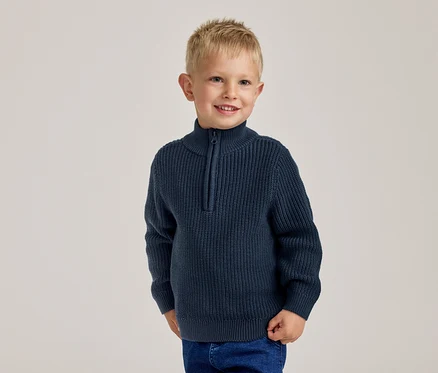 Jeune garçon blond souriant, portant un pull côtelé bleu marine à demi-zip et un jean bleu.
