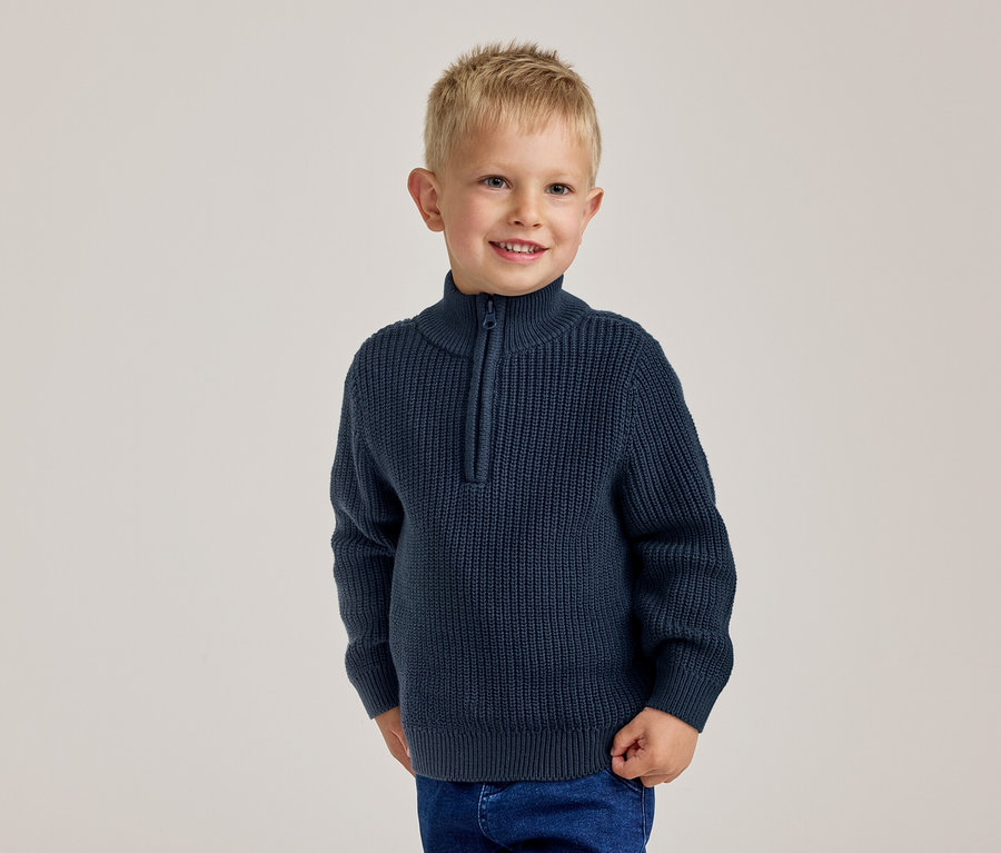 Jeune garçon blond souriant, portant un pull côtelé bleu marine à demi-zip et un jean bleu.