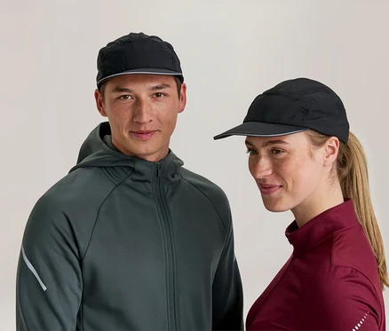 Un homme et une femme portent des casquettes de sport noires. L'homme porte une veste à capuche gris foncé et regarde devant lui, tandis que la femme porte un haut à manches longues bordeaux et sourit légèrement, regardant sur le côté.