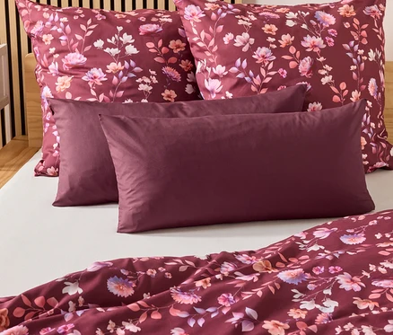 Sur un lit, deux oreillers bordeaux et deux taies d'oreiller à motifs floraux. Une couette bordeaux est posée au pied du lit.