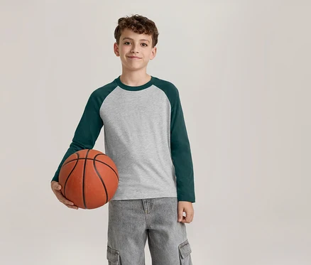 Un garçon souriant vêtu d'un t-shirt à manches longues gris et vert foncé et d'un pantalon cargo gris tient un ballon de basket dans sa main gauche.
