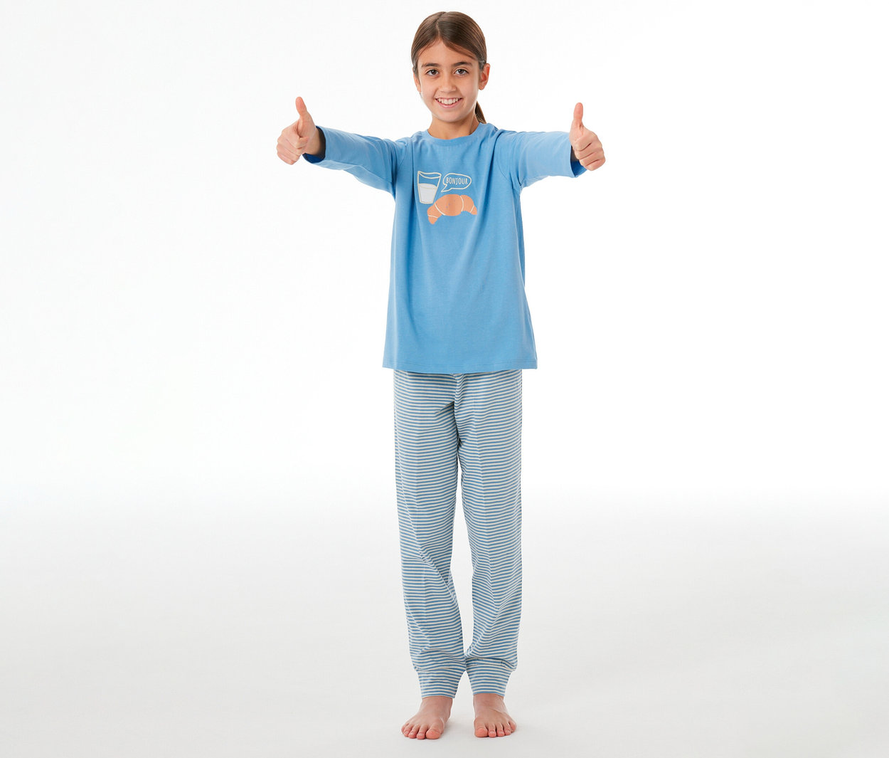 Jeune fille souriante portant un haut de pyjama bleu avec un motif croissant et lait et un bas de pyjama rayé bleu et blanc, levant les deux pouces.