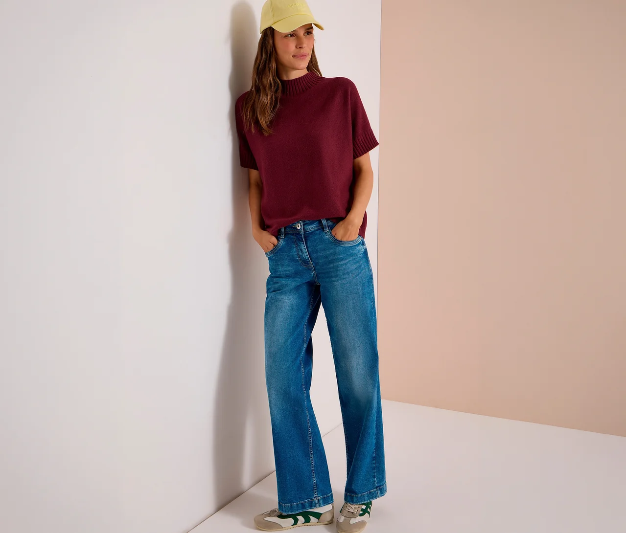 Une femme porte un pull en maille CECIL rouge bordeaux, un jean large et une casquette de baseball jaune, les mains dans les poches, appuyée contre un mur.