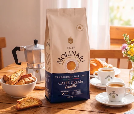 Un paquet de grains de café Caffè Molinari Caffè Crema Gustoso est posé sur une table en bois, à côté d'une cafetière moka argentée, d'un bol de cantuccini, de deux tasses de café, de croissants et d'un vase de fleurs sauvages.