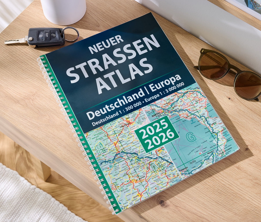 Sur une table en bois sont posés un livre 'Livre	: nouvel atlas routier Allemagne/Europe 2025/2026', une clé de voiture et des lunettes de soleil.