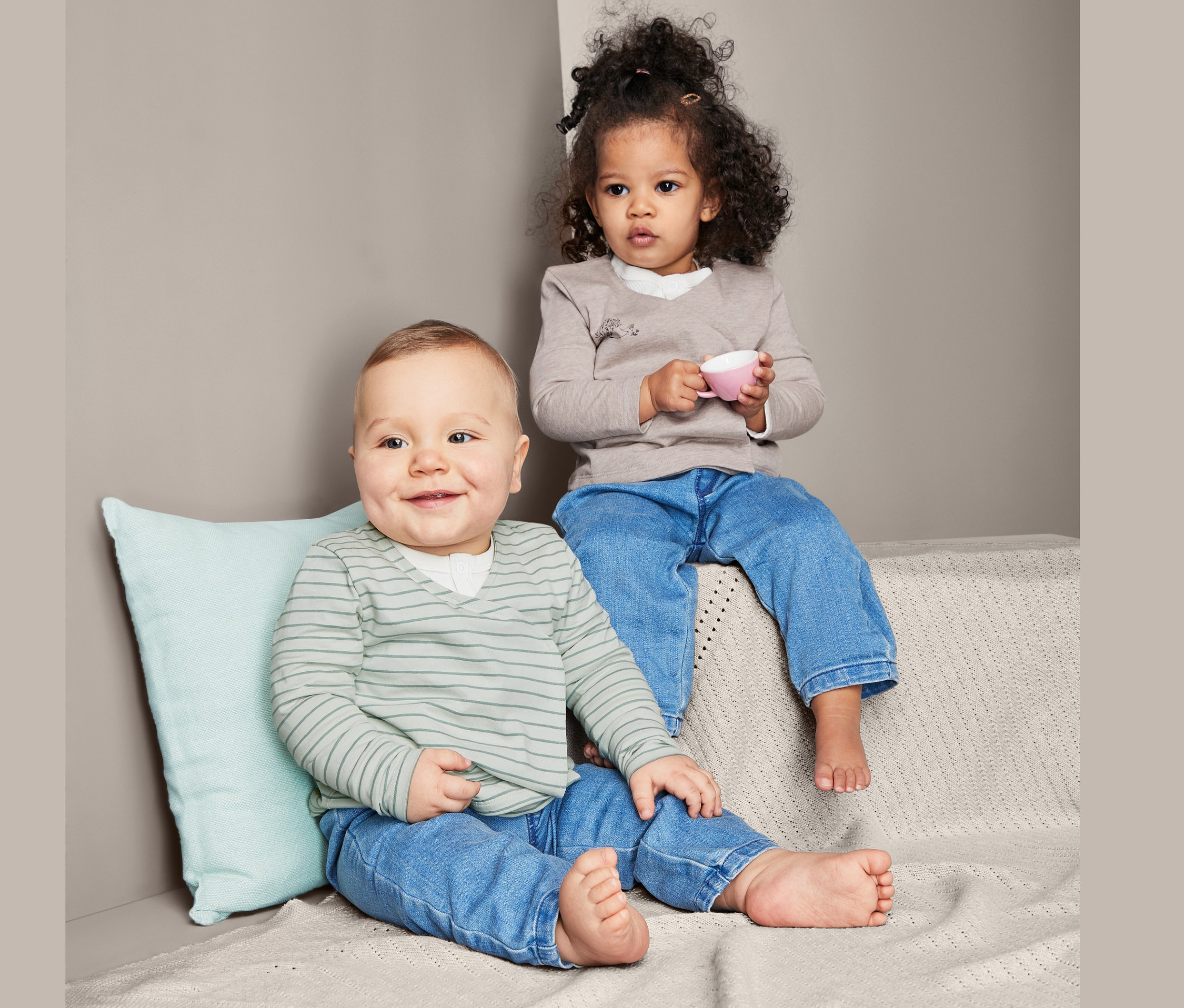 Un bébé est assis à côté d'un enfant en bas âge. L'enfant tient une tasse rose. Ils portent tous les deux un jean et 2 cache-cœurs en jersey de coton.