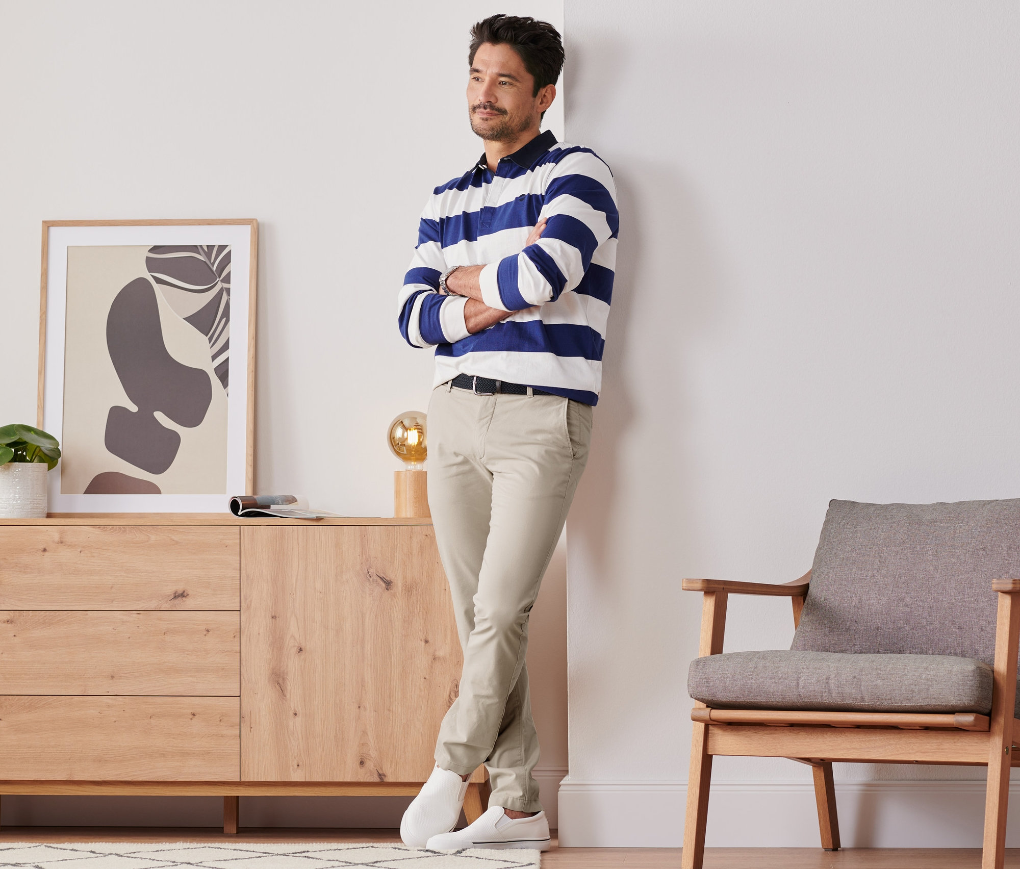 Un homme pose en pantalon chino couleur sable à côté d'une commode et d'une chaise.