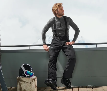 Un homme est assis sur une terrasse en pantalon de ski high-tech, avec un casque et des skis à côté de lui.