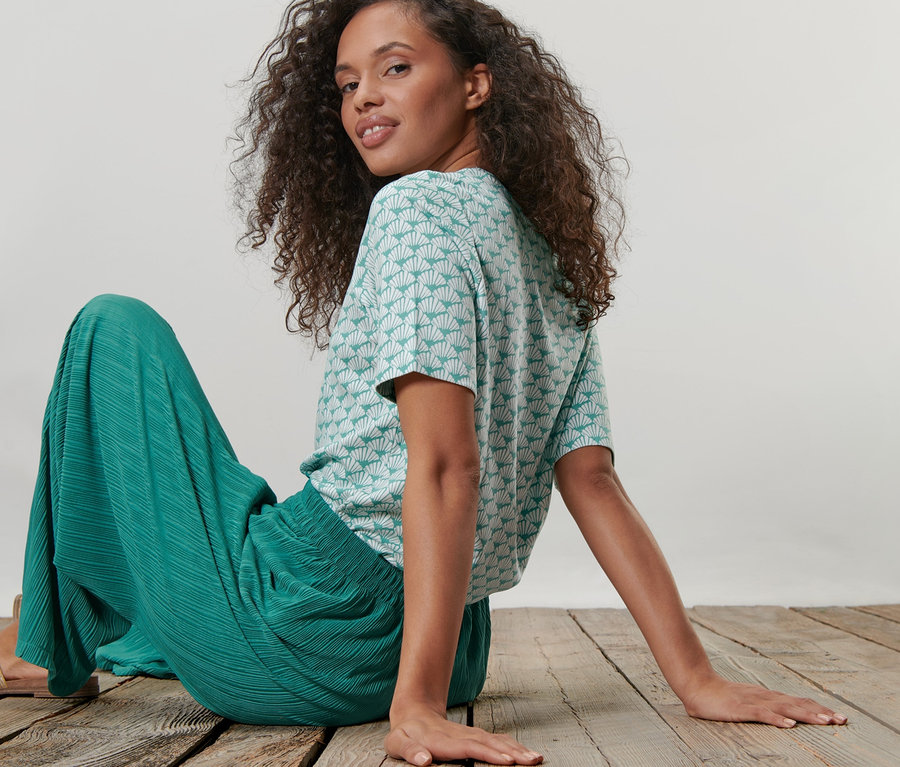 Une femme est assise sur un plancher en bois, portant une chemise en interlock et un pantalon vert.