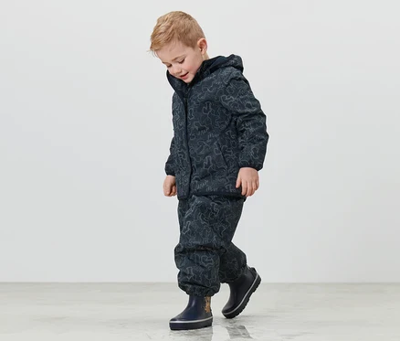 Un garçon portant une veste de pluie thermique pour enfant avec des éléments réfléchissants et des bottes en caoutchouc bleu foncé marche sur le sol.