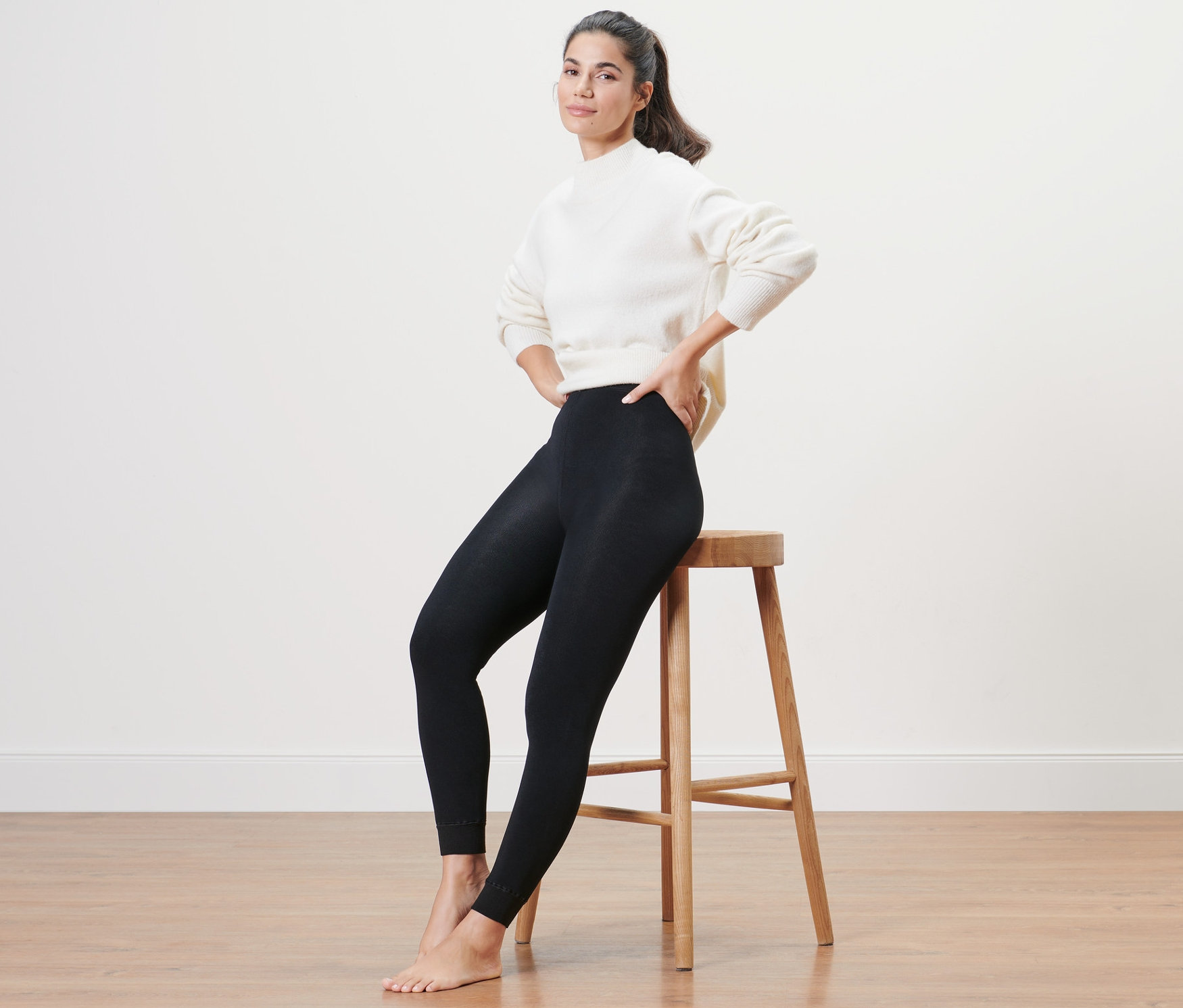 Une femme est assise sur un tabouret et porte un legging doublé et un pull blanc.
