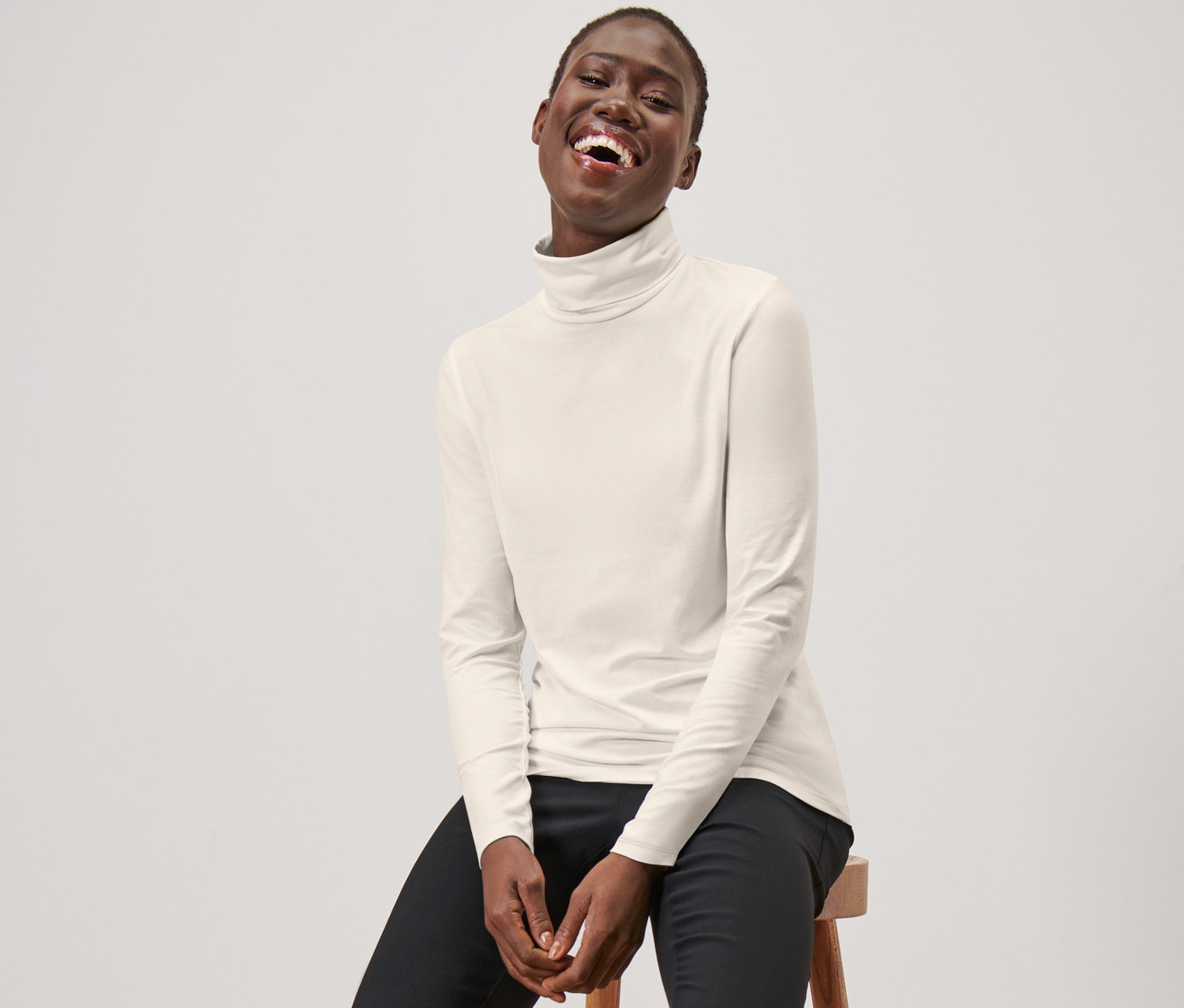 Une femme assise sur un tabouret porte un t-shirt col roulé à manches longues blanc crème, un pantalon noir et des mocassins noirs.