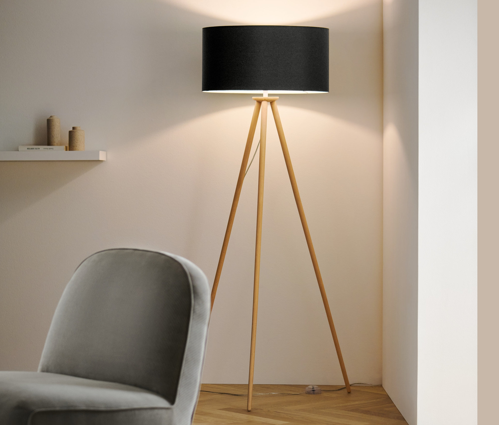 Dans la pièce, un lampadaire en bois « Trépied » avec abat-jour noir et un fauteuil gris.
