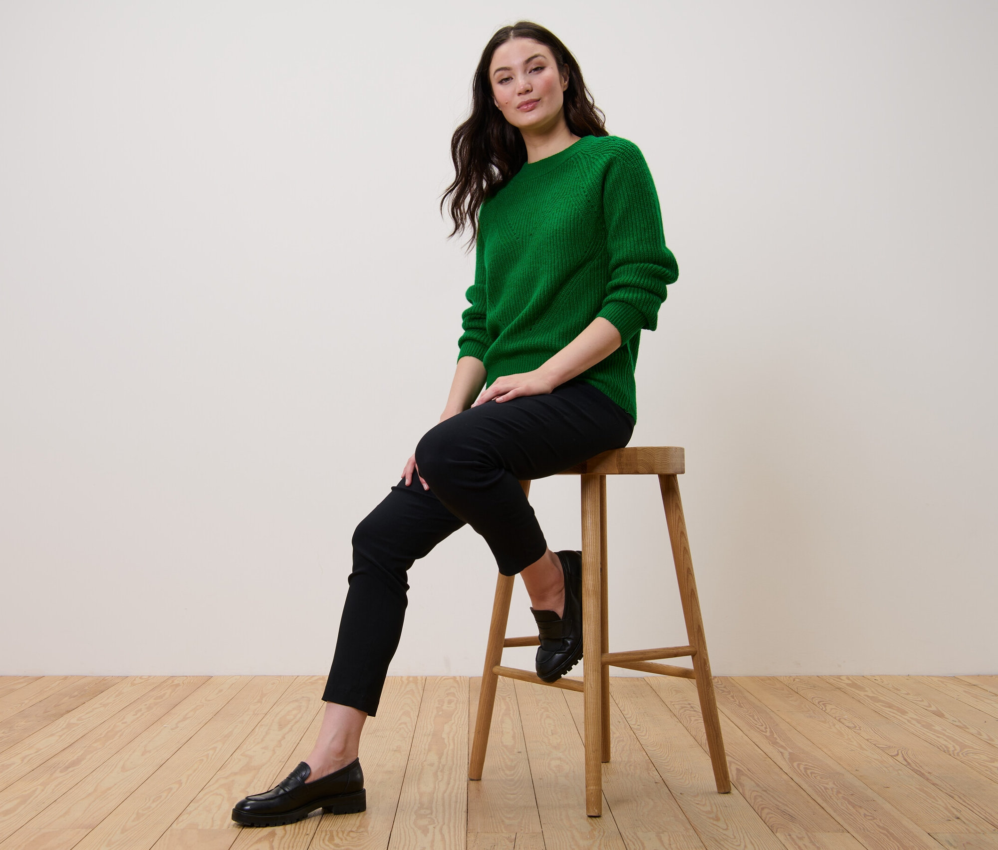 Une femme est assise sur un tabouret et porte un pull en maille vert.