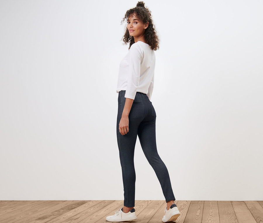 Une femme regardant par-dessus son épaule porte des leggings aspect jeans, un chemisier blanc et des baskets.