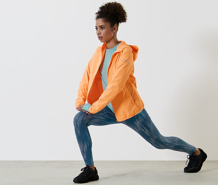 Femme faisant des étirements en veste de course coupe-vent orange fluo, t-shirt technique à manches longues bleu clair et legging de sport réfléchissant argenté.
