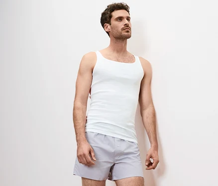 Un homme appuyé contre un mur porte un maillot de corps blanc et un short gris. Le maillot de corps est un 2 maillots de corps finement côtelés.