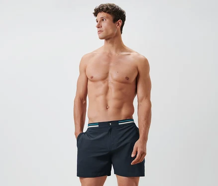 Un homme torse nu pose en short de bain bleu foncé.