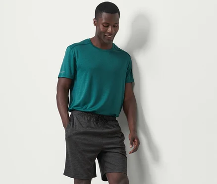 Un homme portant un t-shirt vert et un short de sport anthracite se tient contre un mur blanc.
