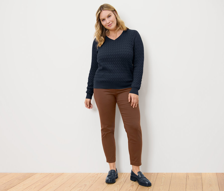 Une femme pose, portant un pull en maille torsadée bleu marine et un pantalon stretch cognac.