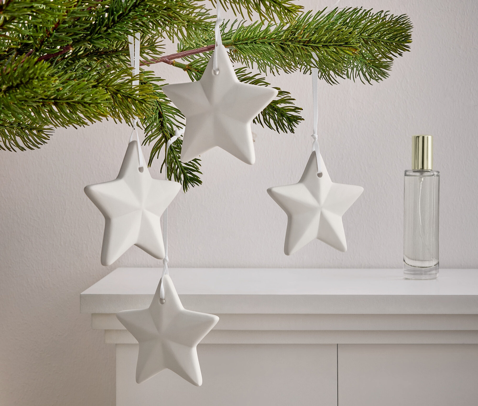 Une branche avec des aiguilles sur laquelle sont suspendues quatre étoiles blanches sur une armoire blanche. A côté des étoiles se trouve un flacon de parfum.