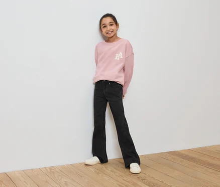 Une fille portant un sweat-shirt rose, un jean noir et des chaussures blanches se tient contre un mur blanc.