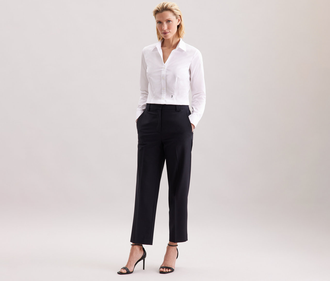 Femme debout portant un chemisier blanc Slim Fit non iron Seidensticker, un pantalon noir et des talons hauts noirs.