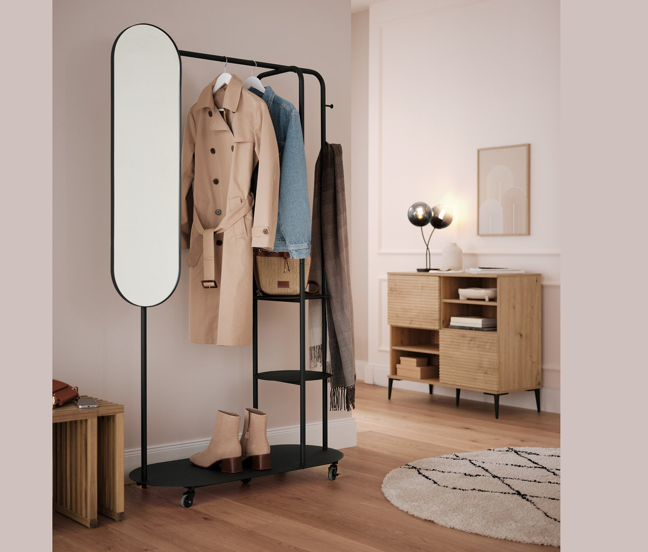 Portant noir avec miroir ovale et tabouret gigogne. Un manteau beige, une veste bleue et une écharpe grise sont suspendus à la barre.