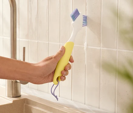 Une main tient une brosse à joints jaune et nettoie les joints entre les carreaux.