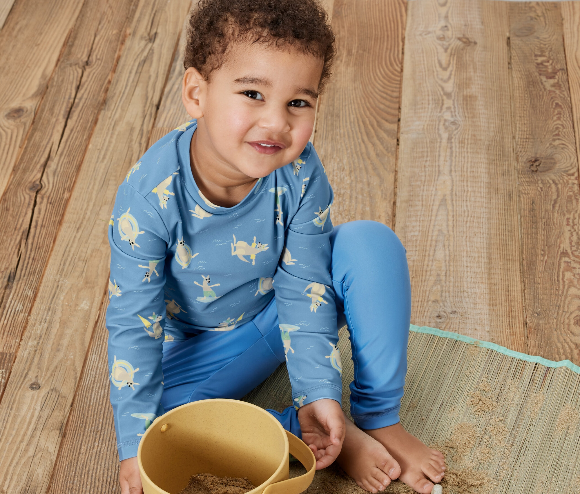 Un garçon est assis sur un plancher en bois et joue avec du sable dans un seau jaune. Il porte un t-shirt pour enfant bleu avec un indice de protection UV 80, kangourou.