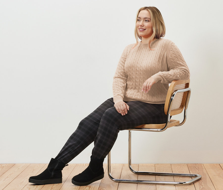 Une femme est assise sur une chaise et porte un pull en maille torsadée crème et un pantalon de jogging à carreaux.