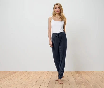 Une femme blonde se tient sur un plancher en bois, portant un débardeur blanc et un pantalon bleu marine.