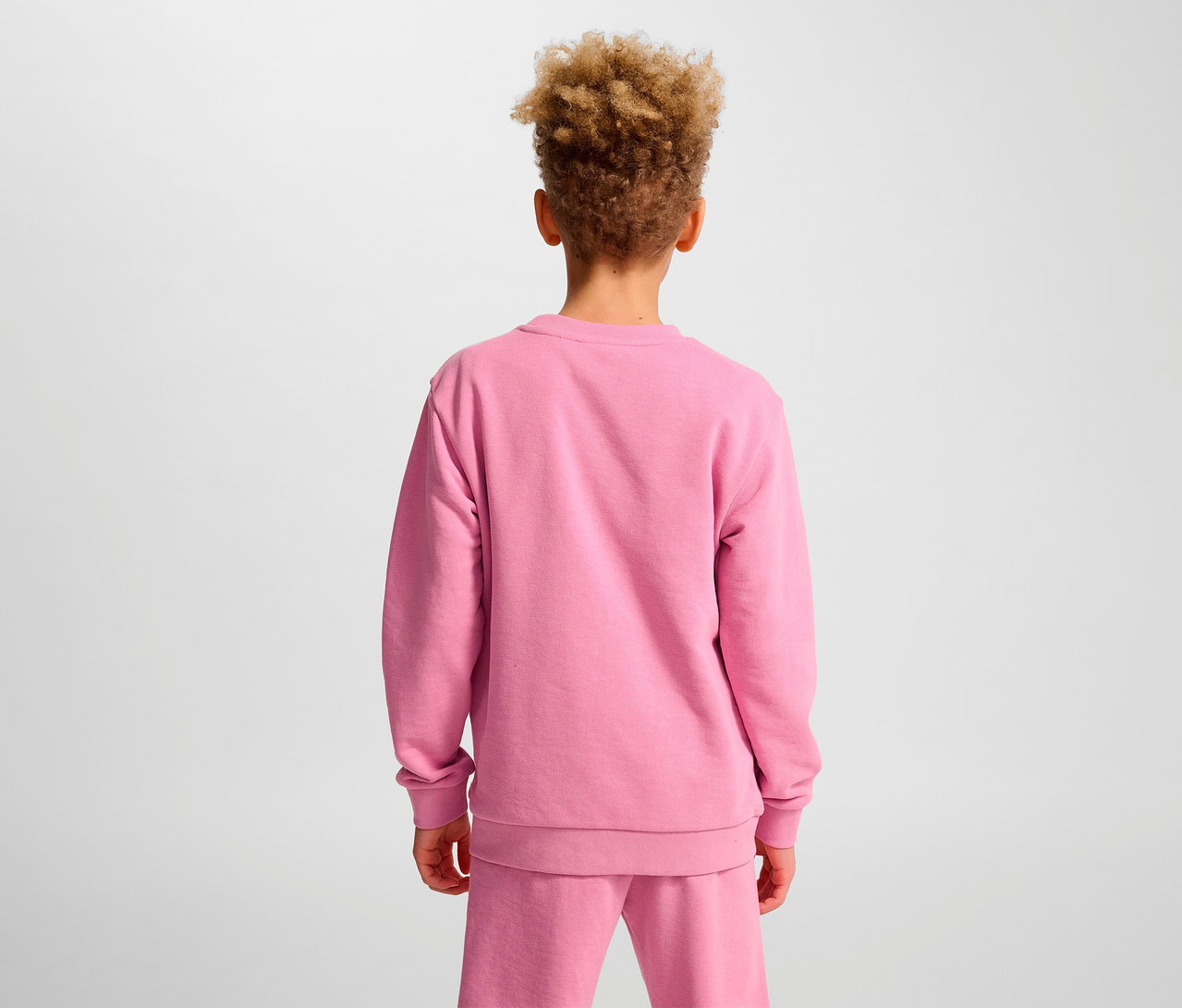 Un garçon aux cheveux bouclés se tient dos à la caméra et porte un sweat-shirt rose HUMMEL HMLJR Base.
