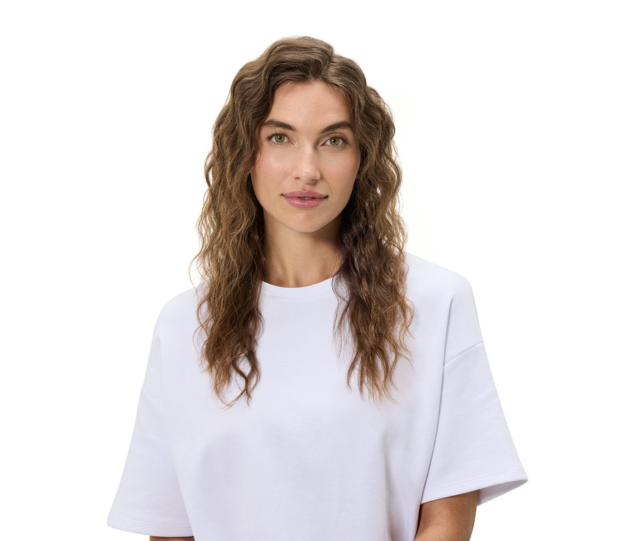 Portrait d'une femme aux cheveux bruns ondulés portant un t-shirt blanc sur un fond blanc.