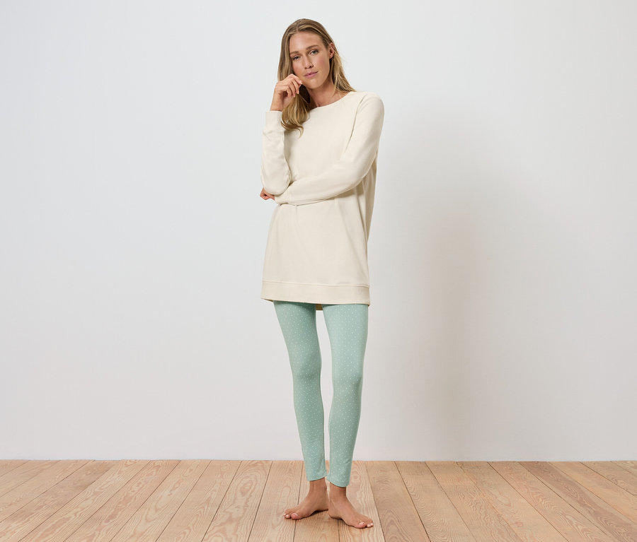 Une femme debout pose dans une tunique crème et des leggings vert clair.