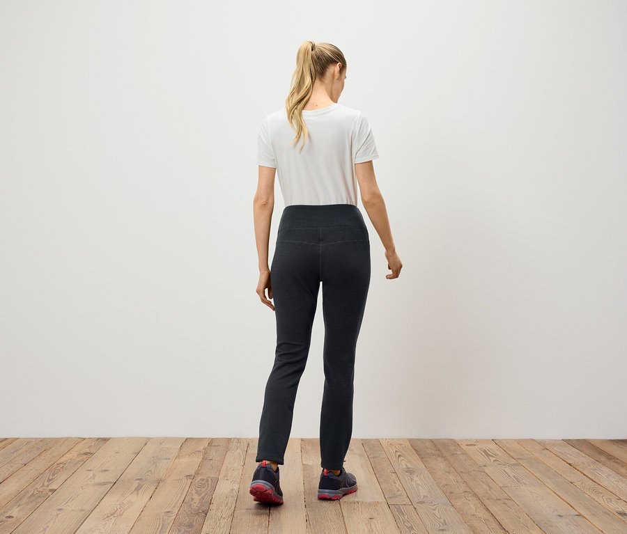 Une femme aux cheveux blonds attachés en queue de cheval se tient dos à la caméra, portant un pantalon noir et un t-shirt blanc.