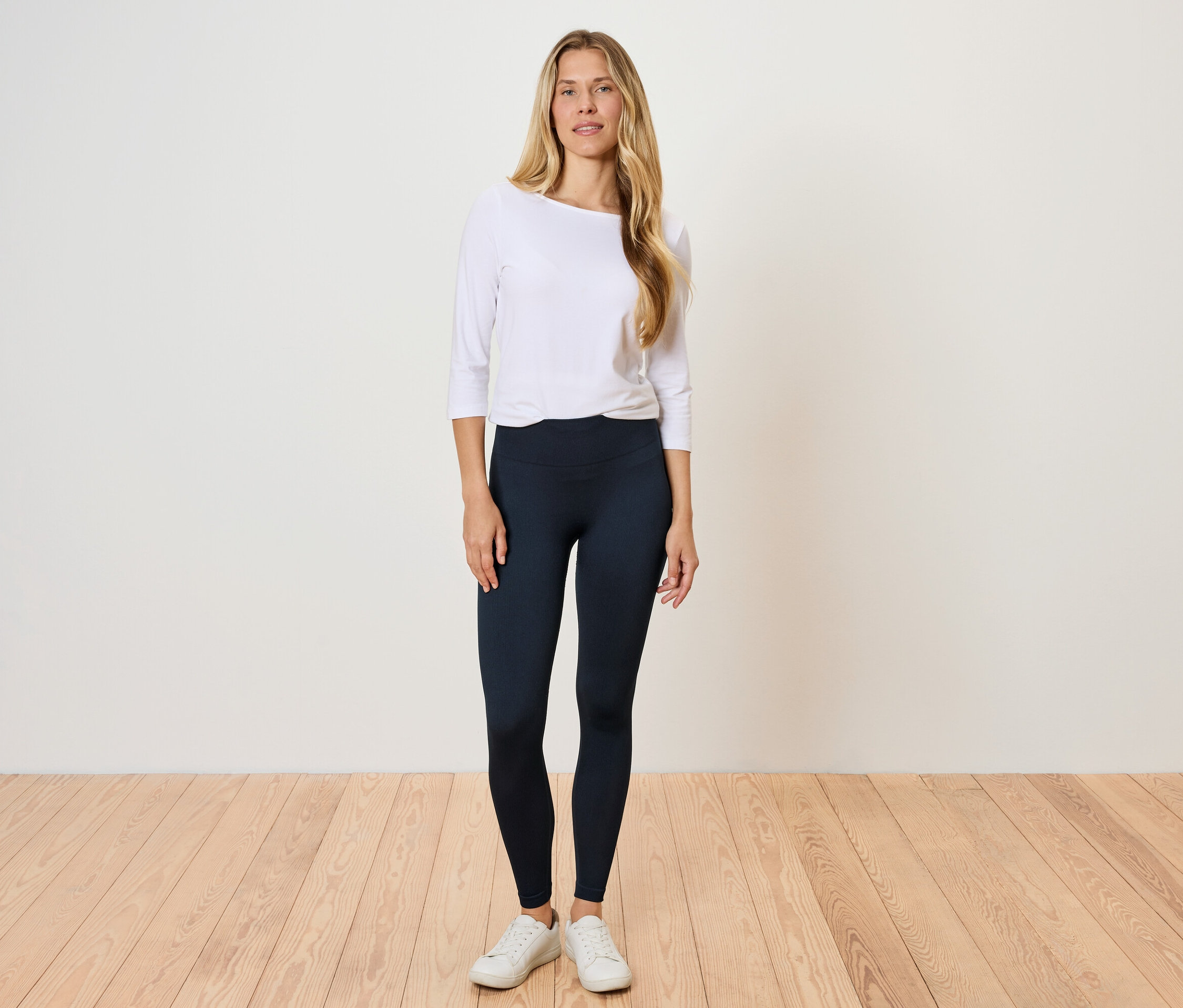 Une femme se tient sur un plancher en bois, portant un t-shirt blanc à manches longues et des leggings noirs.