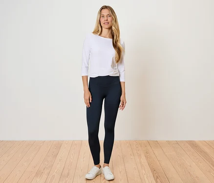 Une femme se tient sur un plancher en bois, portant un t-shirt blanc à manches longues et des leggings noirs.