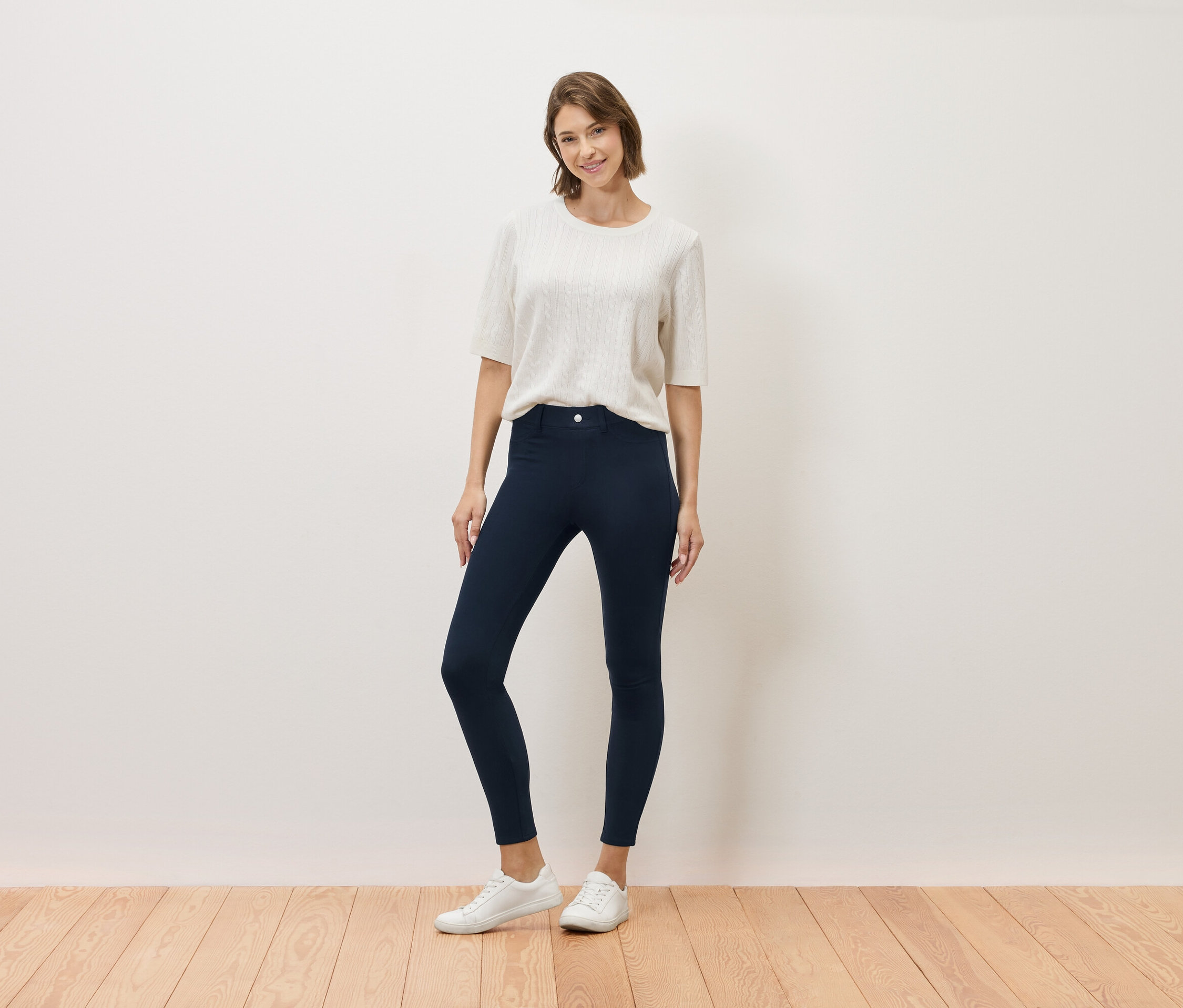 Une femme pose, portant un haut couleur crème, un pantalon bleu marine et des baskets blanches.