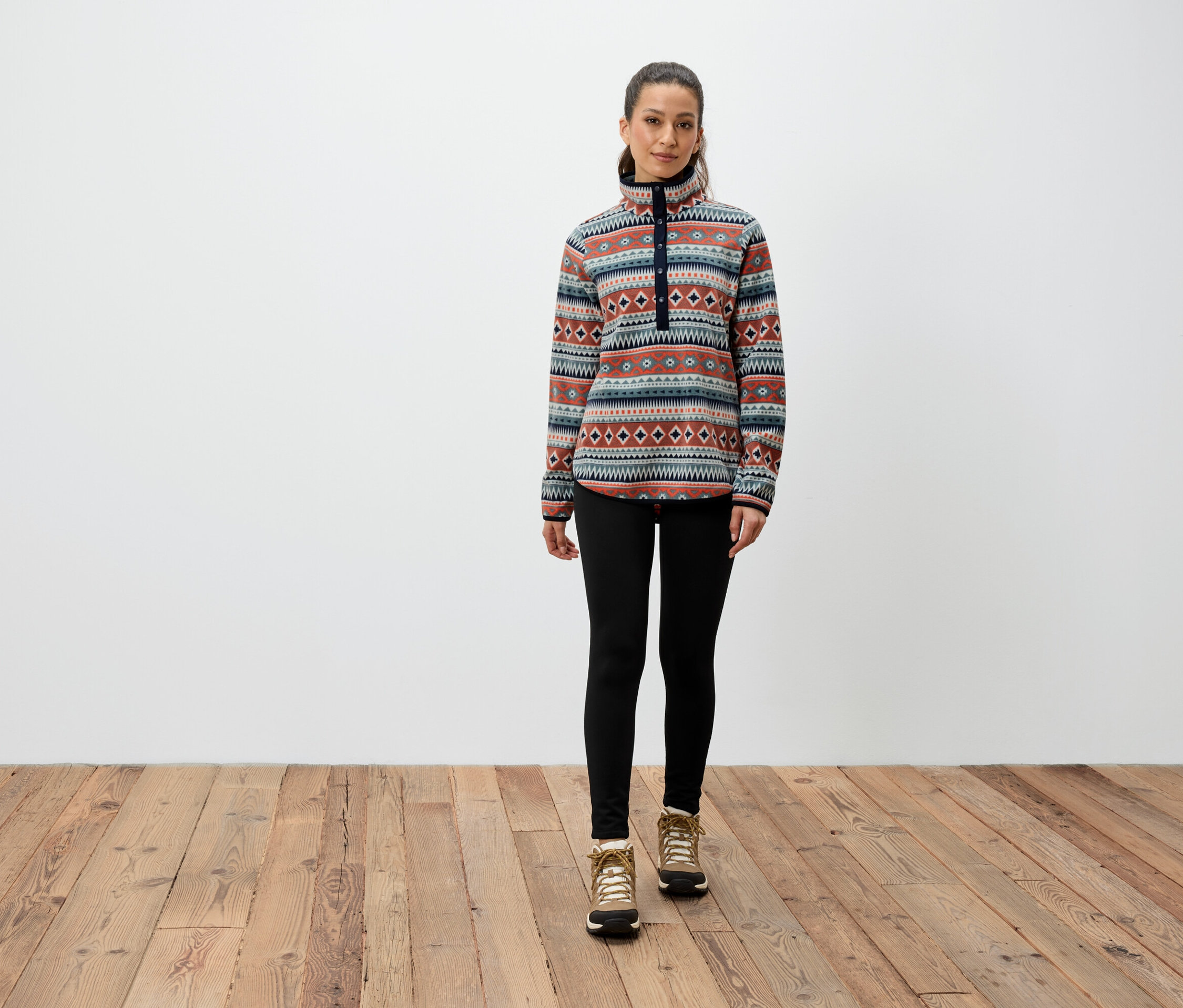 Une femme se tient sur un plancher en bois et porte un sweat-shirt à motifs, des leggings noirs et des bottes marron.