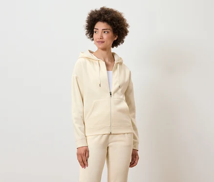 Une femme aux cheveux bouclés pose dans un sweat à capuche zippé de couleur crème et un pantalon de survêtement.