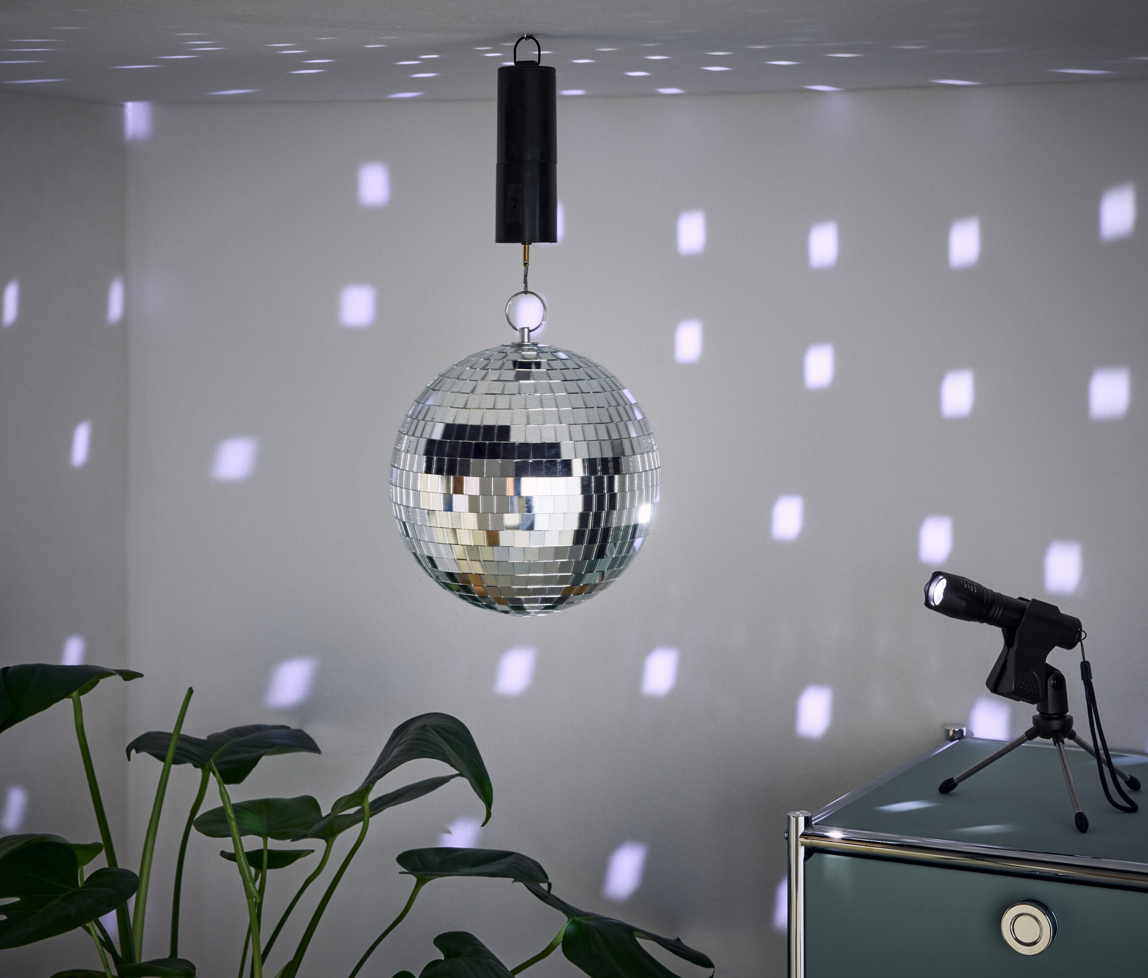 Une boule à facettes est suspendue au plafond, projetant des taches de lumière sur les murs. Un projecteur est posé sur un meuble à côté et une plante est au premier plan.
