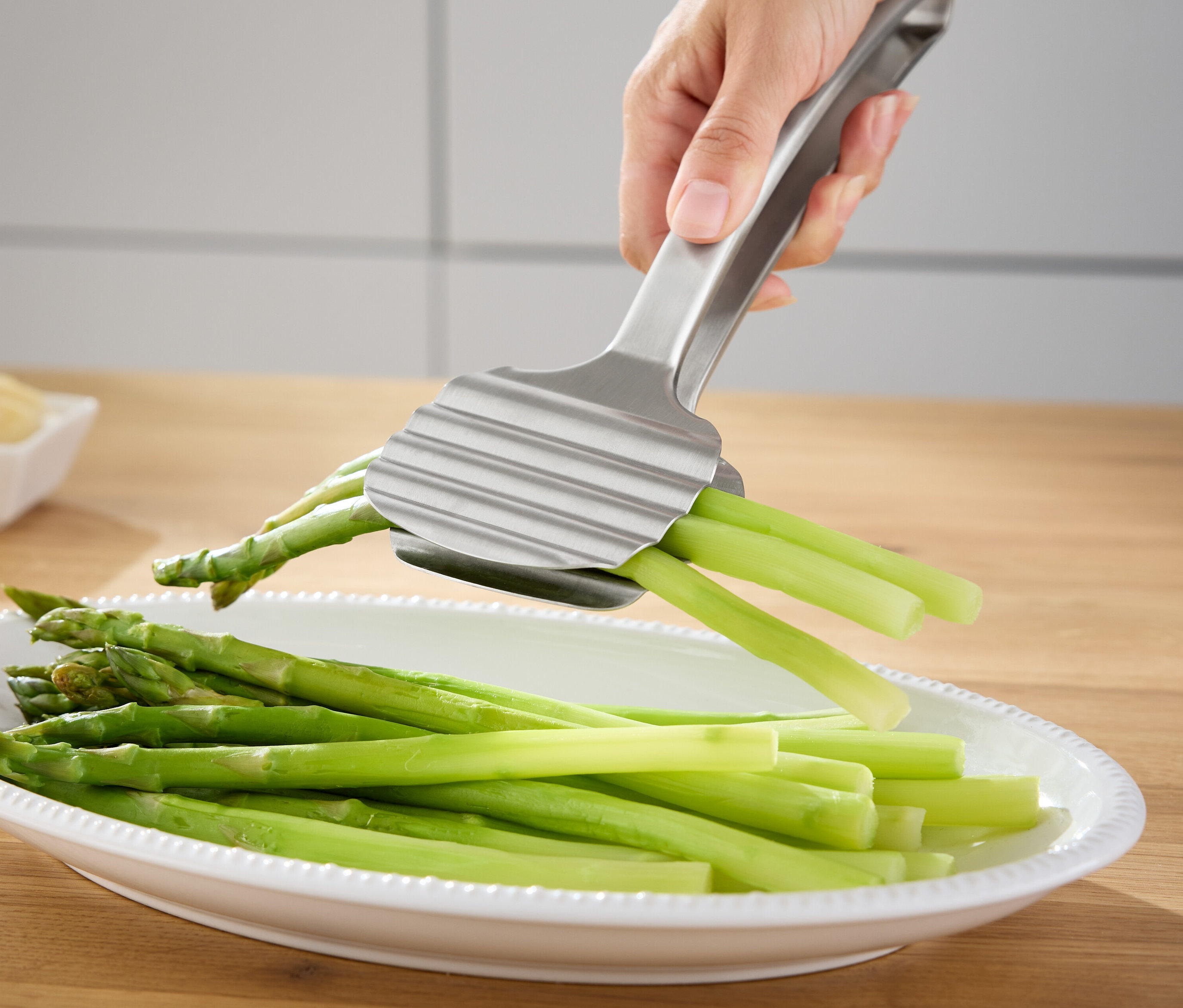 Une main tient une pince à asperges argentée au-dessus d'un plat d'asperges vertes.