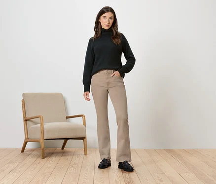 Femme portant un pull à col roulé noir et un pantalon beige posant à côté d'un fauteuil beige.