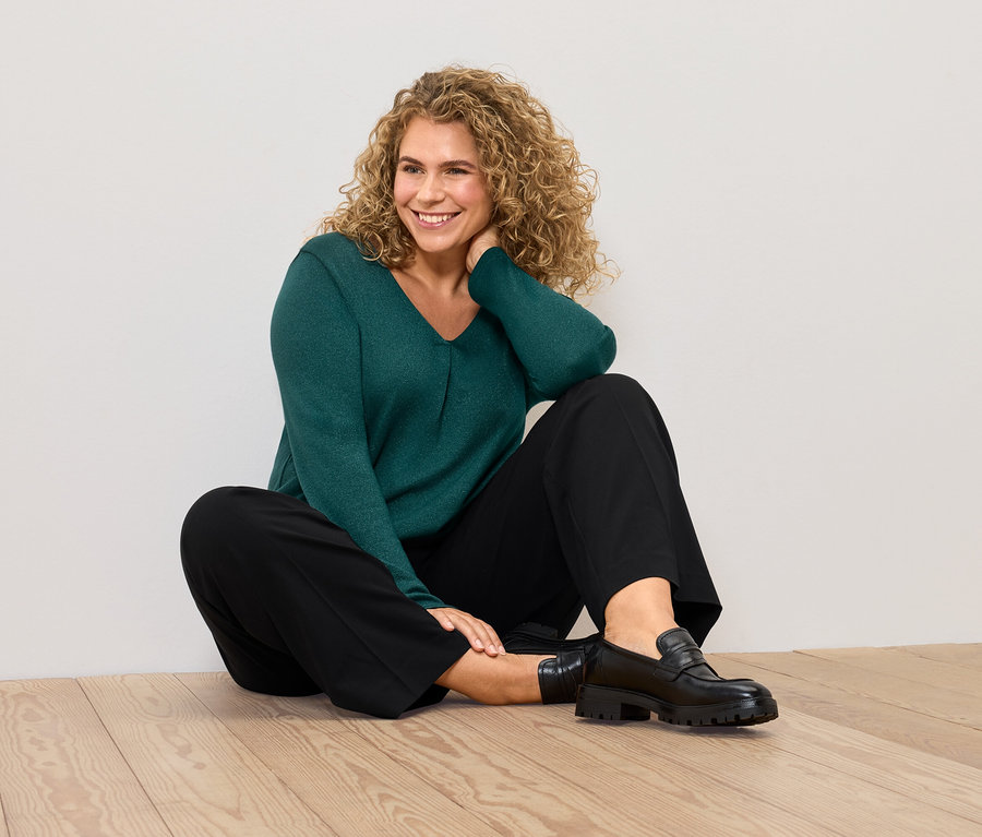 Une femme est assise par terre, portant un pull vert et un pantalon noir.